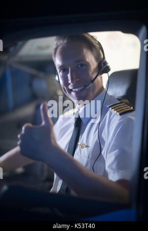 Portrait of smiling young male pilot showing thumbs up while sitting in airplane cockpit Stock Photo