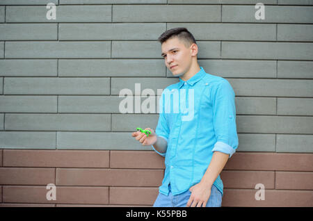Pensive young man on the azure shirt  and blue jeans hand holding popular gadget fidget spinner. Teenager playing with green spinner outdoors  on the  Stock Photo