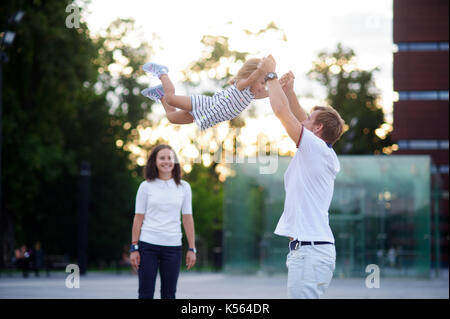 Young family on walk. The father cheerfully plays with the little daughter. He throws up his baby. Mother stands and smiles. Stock Photo
