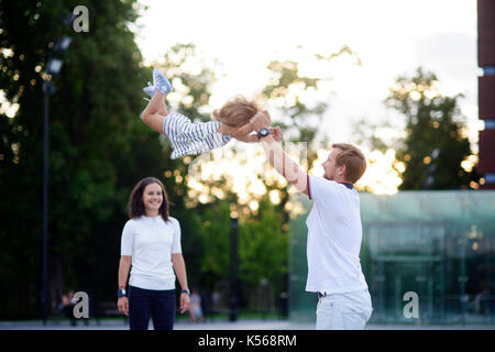 Young family on walk. The father cheerfully plays with the little daughter. He throws up his baby. Mother stands and smiles. Stock Photo