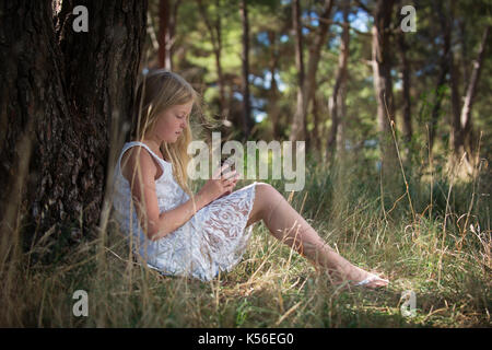 Long blonde haired cute girl in white dress sitting leaning her back against tree looking at pine cone in her hands. Stock Photo
