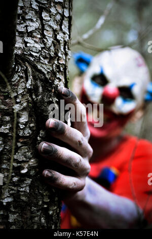 closeup of a scary evil clown wearing a dirty red costume with a creepy smile in his face while is hiding behind a tree in the woods Stock Photo