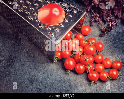 Preparation recipe tomato juice. Large tomato and old grater down to small grape cherry tomatoes on retro vintage rustic gray stone background. Stock Photo