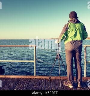 Travel photographer on pier. Photo shooting during sunny autumnal afternoon at handrail on the wooden mole. Stock Photo