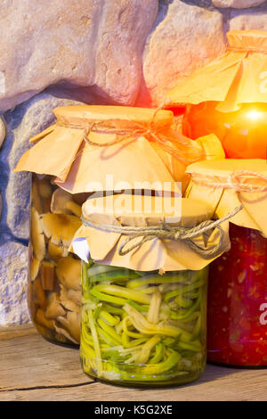 Home canned vegetables in glass jars near the stone wall. Pickled ramsons, mushrooms, tomato sauce. The concept of natural product preparations for th Stock Photo