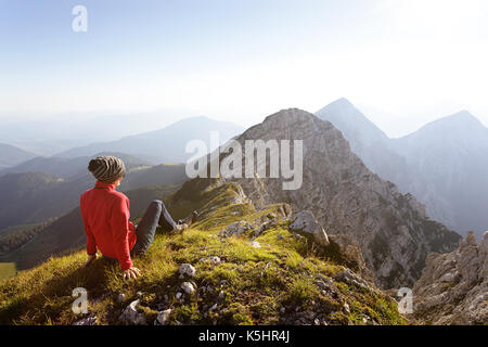 Woman in red hiking jacket sitting on grass on top of the mountain with stunning view over the peaks. Kladivo, Karakvanke, Slovenia. Stock Photo
