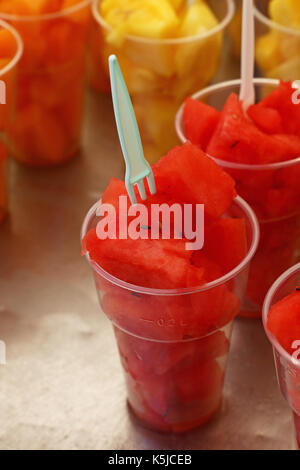 Street food, selection of fruit salads, slices of cut fresh ripe watermelon and melon cubes in plastic cups with forks at retail market stall display, Stock Photo