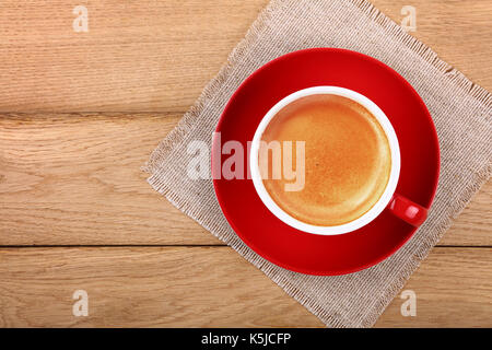 Full big cup of espresso coffee with brown crema froth on red porcelain saucer over wooden table with textile tablecloth napkin, close up, elevated to Stock Photo