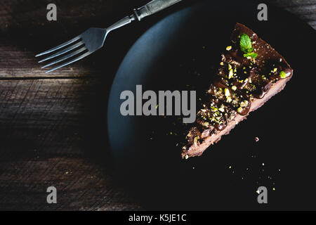 Dark chocolate cheesecake decorated with chopped pistachios and mint leaf on black plate on wooden table. Top view, toned image Stock Photo