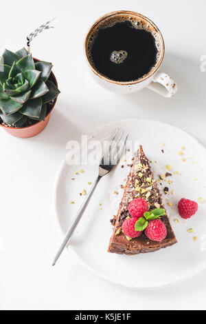 Slice of chocolate cake with raspberries and nuts, cup of black coffee and succulent plant stone rose on white table. Feminine breakfast food photogra Stock Photo