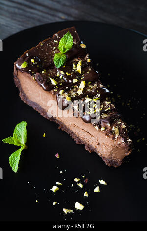 Dark chocolate cheesecake decorated with chopped pistachios and mint leaf on black plate on wooden table. Closeup view Stock Photo
