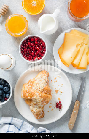 Continental breakfast table top view. Fresh croissants, boiled egg, cheese, orange, jam, honey, cream, blueberries and currants on bright blue table.  Stock Photo