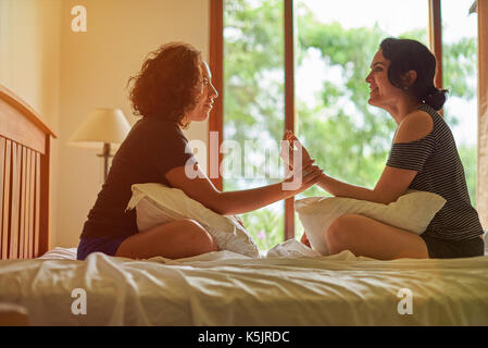 Two hispanic woman talk on bed in room with big window Stock Photo