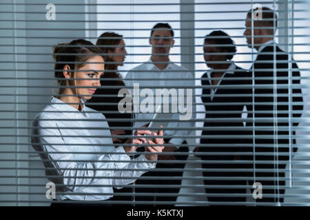 Young Businesswoman With Digital Tablet Standing Behind Blind In Office Stock Photo