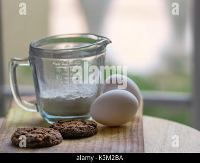 A glass measurement cup, two eggs and baked brown chocolate cookies on top of a wooden board, with soft light and out of focus background Stock Photo
