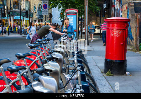 A man and woman try and work out how to hire a Santander bike form the docking station on Castlehaven Road in Camden Town, London. Stock Photo