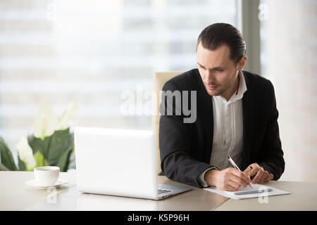 Young businessman in suit looking at laptop screen, taking notes Stock Photo