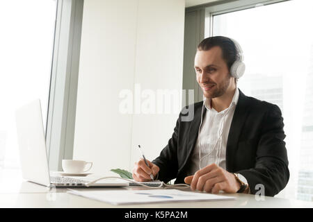 Businessman in headphones taking notes in front of laptop. Stock Photo