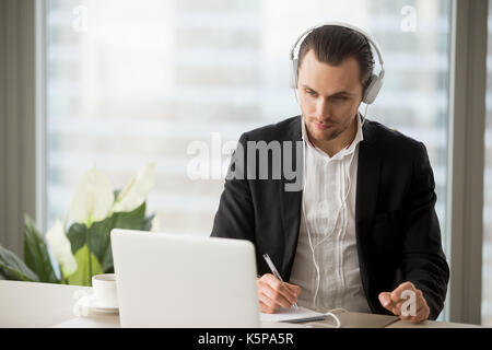 Businessman in headphones taking notes in front of laptop. Stock Photo