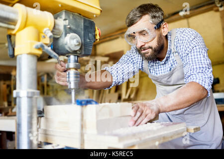 Handsome bearded carpenter wearing safety goggles and apron using drill press machine in order to make holes in wooden plank, interior of spacious wor Stock Photo