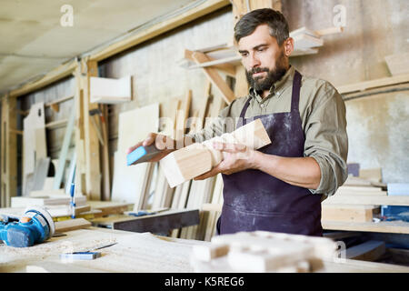 Handsome bearded craftsman using sandpaper in order to remove paint from wooden detail, interior of spacious workshop on background Stock Photo
