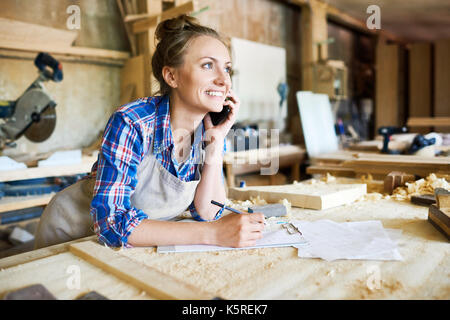 Cheerful young carpenter discussing order details with her customer on smartphone and taking necessary notes, interior of spacious workshop on backgro Stock Photo