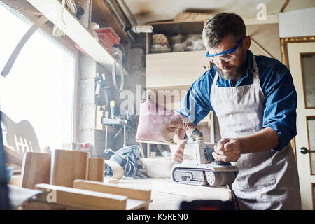 Confident bearded carpenter wearing apron and safety goggles working with belt sander, interior of messy workshop on background Stock Photo