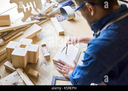 Over shoulder view of experienced carpenter using compass in order to draw circle on wooden plank, messy table covered with sawdust Stock Photo
