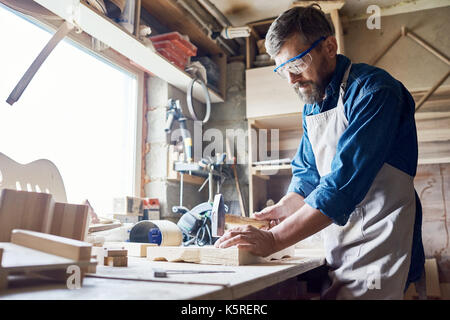 Bearded middle-aged craftsman wearing safety goggles and apron hammering nails into board, interior of messy workshop on background Stock Photo