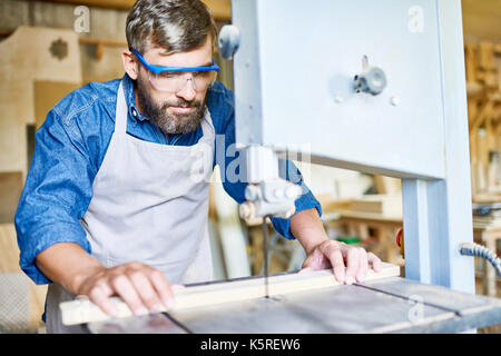 Middle-aged bearded craftsman wearing apron and safety goggles using sawing machine while working with wooden plank, blurred background Stock Photo