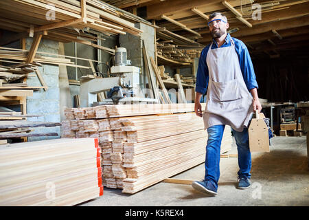 Full length portrait of confident bearded carpenter wearing apron and safety goggles walking along workshop with tools kit in hand Stock Photo