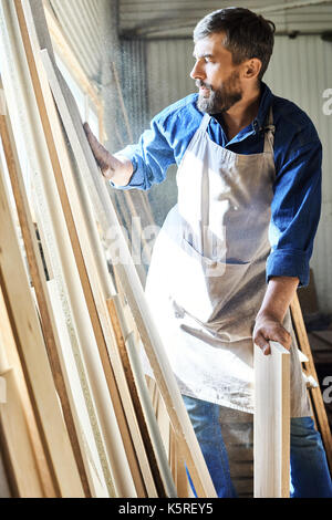 Handsome bearded carpenter wearing denim shirt and apron evaluating quality of wooden board while wrapped up in work at spacious workshop Stock Photo