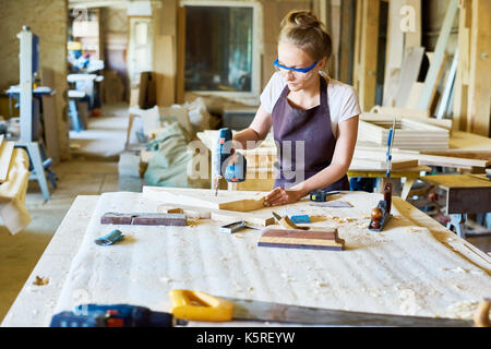 Attractive fair-haired carpenter in safety goggles and apron making holes in wooden detail with help of electric drill, interior of spacious workshop  Stock Photo