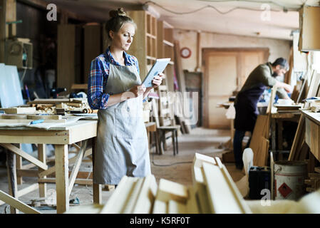 Full length portrait of concentrated young craftswoman in apron standing at spacious workshop and using digital tablet Stock Photo