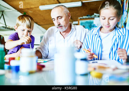 Mature drawing teacher giving lesson to his little pupils while sitting at desk in spacious art studio Stock Photo