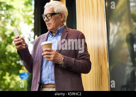 Portrait of modern senior man using smartphone outdoors in city, holding coffee cup Stock Photo