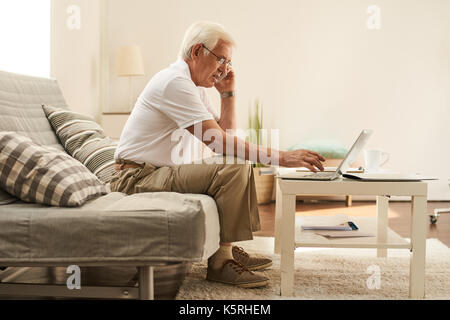 Side view portrait of modern senior man using laptop at home and speaking by phone Stock Photo