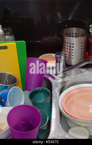 A collection of dirty dishes which includes plates, bowls, glasses, cutlery & chopping boards, stacked in a dirty kitchen sink with mixer tap. Stock Photo