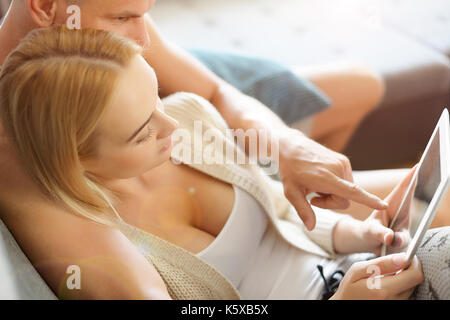 Young stylish couple cuddling on the sofa using digital tablet at their modern home. Woman holding tablet, browsing the web. Stock Photo