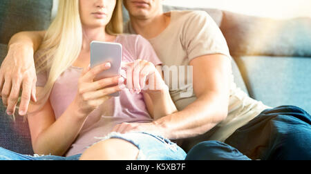 Young unrecognisable couple cuddling on the sofa checking smartphone at their modern home. Woman holding mobile phone, browsing the web. Stock Photo
