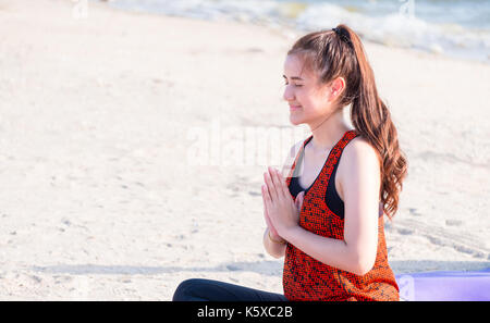 asian woman seated doing hand namaste yoga pose on sand at coastline beach, Calm and relax concept,wellness and healthy lifestyle. Stock Photo