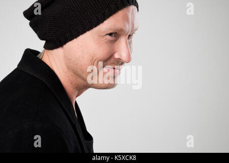 Smiling young man in black clothes standing and hat against white background looking aside Stock Photo