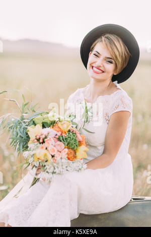 The smiling bride with the big colourful bouquet sitting on the vintage suitcase among the sunny field. Stock Photo