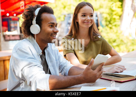 Side view of two smiling young friends sitting by the table outdoors and using smartphone Stock Photo