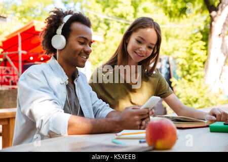 Side view of two pleased young friends sitting by the table outdoors and using smartphone Stock Photo