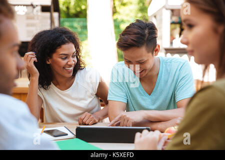Two friends sitting outdoors by the table with their friends and using laptop computer Stock Photo