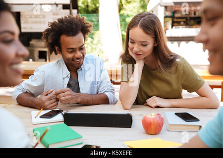 Two young smiling friends sitting outdoors by the table with their friends and using laptop computer Stock Photo