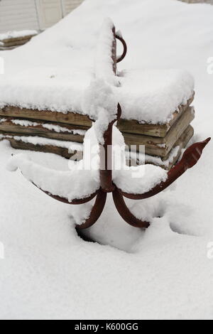 Old rusty grapnel anchor with five tines left aside on the snow covered ground beside a pile of logs in the courtyard of a white cottage-tourist rorbu Stock Photo