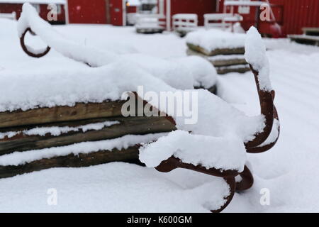 Old rusty grapnel anchor with five tines left aside on the snow covered ground beside a pile of logs in the courtyard of a white cottage-tourist rorbu Stock Photo