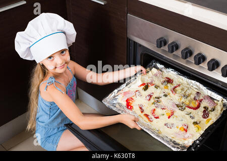 Smiling russian girl in cook cap near oven with pizza Stock Photo